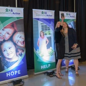 Two women taking a selfie with their cellphone in front of Community Action banners that are illuminated with uplighting