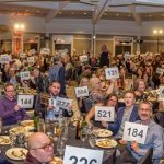 Room full of happy and excited guests sitting around dinner tables while eating and raising bid paddles